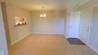 This single-floor space presents a clean, functional layout centered around a generously sized living area finished with wall-to-wall beige carpeting that adds warmth underfoot. The room is anchored by a classic multi-arm chandelier suspended from the ceiling, providing both ambient illumination and a subtle decorative focal point. Neutral, warm-toned walls create a calm and versatile backdrop throughout the space.

A notable architectural feature is the pass-through opening on the left wall, which connects the living area to the kitchen. This cut-out is framed with cabinetry above and features what appears to be a stone or granite countertop ledge below, offering both visual connectivity between the two spaces and practical utility as a serving or bar-style surface.

A white interior door on the right side of the living area leads to an adjacent room or utility space, transitioning from the carpeted living area to dark hardwood or laminate flooring beyond the threshold. White baseboards throughout provide crisp contrast against the neutral walls and flooring. The overall ambiance is bright, tidy, and move-in ready, well-suited for a variety of furnishing styles.