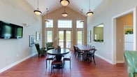 Jackson Green is a thoughtfully designed single-floor communal gathering space that combines classic architectural details with a warm, welcoming atmosphere. The centerpiece of the space is a grand main room featuring soaring vaulted ceilings adorned with elegant pendant lighting, creating an airy and open environment. Expansive multi-pane windows and French doors line the walls, flooding the interior with natural light while offering views of the surrounding greenery outdoors.

Rich hardwood flooring in a warm reddish-brown tone extends throughout, grounding the space with a sense of comfort and sophistication. The layout is thoughtfully divided into distinct functional zones. A lounge area features plush green wingback chairs arranged around a small accent table, accented by framed black-and-white nature photography on the walls. A separate seating area offers a neutral sofa, wall-mounted television, and glass coffee table for relaxed gatherings.

The dining area is anchored by a substantial wooden dining table surrounded by classic black bentwood chairs, accommodating a sizable group. A pass-through window connects this area to an adjoining kitchen, adding practicality for events and catering. The overall color palette of soft greens, warm beiges, and neutral tones contributes to a calm and inviting ambiance suited for community gatherings and social interaction.
