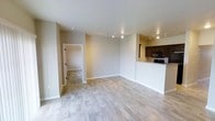 This single-floor space is thoughtfully designed with an open-concept layout that seamlessly connects the main living area to the kitchen. Light-toned hardwood flooring runs throughout the living space, lending warmth and a clean, contemporary aesthetic to the interior.

The living area is generously proportioned, featuring neutral beige walls and white baseboards that create a bright, welcoming atmosphere. Track lighting is installed along the ceiling, providing flexible and functional illumination throughout the space.

The kitchen is partially separated by a raised breakfast bar counter finished with granite countertops, offering both a casual dining option and a natural visual boundary between spaces. Dark espresso-toned cabinetry lines the kitchen walls, complemented by black appliances, including a built-in microwave, creating a bold contrast against the lighter tones of the living area.

An adjacent room, visible through an open doorway, features tile flooring, suggesting a utility, bathroom, or entryway space. Overall, the unit presents a well-balanced blend of modern finishes and practical design elements suited for comfortable everyday living.