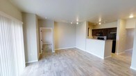 This single-floor space is thoughtfully designed with an open-concept layout that seamlessly connects the main living area to the kitchen. Light-toned hardwood flooring runs throughout the living space, lending warmth and a clean, contemporary aesthetic to the interior.

The living area is generously proportioned, featuring neutral beige walls and white baseboards that create a bright, welcoming atmosphere. Track lighting is installed along the ceiling, providing flexible and functional illumination throughout the space.

The kitchen is partially separated by a raised breakfast bar counter finished with granite countertops, offering both a casual dining option and a natural visual boundary between spaces. Dark espresso-toned cabinetry lines the kitchen walls, complemented by black appliances, including a built-in microwave, creating a bold contrast against the lighter tones of the living area.

An adjacent room, visible through an open doorway, features tile flooring, suggesting a utility, bathroom, or entryway space. Overall, the unit presents a well-balanced blend of modern finishes and practical design elements suited for comfortable everyday living.