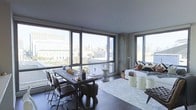 This single-floor space showcases a seamlessly integrated open-plan layout that combines a dining area and a living lounge within one expansive, light-filled interior. Defining the space are dramatic floor-to-ceiling windows that span nearly the entire exterior wall, framing panoramic views of the surrounding urban skyline. The abundant natural light these windows provide enhances the airy, contemporary atmosphere throughout.

The dining area is anchored by a substantial dark wood table paired with sleek, dark leather chairs, offering a refined yet understated setting for meals. Adjacent to it, the living area features a comfortable, neutral-toned sofa dressed with accent cushions, positioned atop a soft area rug. A collection of cylindrical side tables in muted tones adds functional versatility to the lounge space.

The overall design aesthetic leans industrial-modern, characterized by a cool, neutral color palette of grays and dark tones, concrete-finish ceilings, and dark hardwood flooring. The interplay of raw materials and curated furnishings creates a sophisticated, urban ambiance. The corner window configuration further maximizes the cityscape views, making the outdoor environment a central visual feature of the interior.