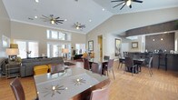 This single-story open-plan space is designed to accommodate both relaxed lounging and casual dining within a seamlessly connected layout. The most striking architectural feature is the soaring vaulted ceiling, which lends an expansive, airy quality to the entire interior. Multiple modern ceiling fans are suspended at varying heights, ensuring comfort throughout the space while adding a contemporary visual element.

The flooring is warm hardwood, running continuously across the room and tying the different functional zones together cohesively. The dining area features multiple square glass-topped tables surrounded by dark leather chairs, offering a polished yet inviting setting. A second dining cluster with a darker, more intimate aesthetic is positioned to the right, set apart by a curved accent wall with rich stone-like detailing.

The lounge area, situated toward the back, includes comfortable upholstered sofas and accent seating in earthy tones, anchored by large windows that draw in abundant natural light. Recessed lighting complements the natural illumination throughout. Framed artwork on the walls and decorative table lamps add warmth, creating a welcoming and well-appointed communal atmosphere.