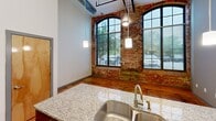 This loft-style residence at 1101 Hawthorne Lane showcases a striking industrial aesthetic rooted in its original architecture. Soaring ceilings with exposed ductwork and dramatic arched windows framed by original brick walls flood the living area with natural light and outdoor views. Warm hardwood floors run throughout, complementing the open-concept layout. The well-appointed kitchen features rich dark cabinetry, granite countertops, a large island with undermount sink, stainless steel appliances, and stylish pendant lighting. The bedroom offers a wood sliding barn door and built-in closet storage. The bathroom continues the granite theme with an undermount sink and full mirror vanity. A true urban retreat blending historic character with modern finishes.