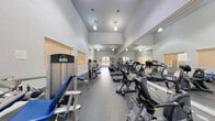 Located at 550 NW 61st St in Coconut Creek, Florida, this fitness center offers a well-appointed, single-floor exercise space designed to accommodate a comprehensive workout experience. The room features soaring ceilings that lend an open, airy atmosphere, enhanced by a row of pendant light fixtures that provide consistent illumination throughout. Large arched windows along one wall invite abundant natural light and offer views of the surrounding tropical greenery, creating an inviting environment for exercise. A full-length wall mirror runs along one side of the room, visually expanding the space and allowing users to monitor their form during workouts. The flooring consists of durable rubber gym matting, providing safety and comfort underfoot. The facility is equipped with a varied selection of cardio machines, including treadmills, ellipticals, stationary bikes, and rowing machines, all arranged in an organized row for easy access. A dedicated free weights area features a multi-tiered dumbbell rack with a range of weights, an adjustable workout bench, and a cable resistance machine for targeted strength training. Additional strength training machines are positioned throughout the space. Access to the fitness center is provided through elegant glass-paned French doors, contributing to the refined architectural character of the facility.