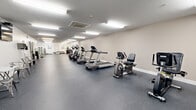 Located at 233 Lake Ave in Saratoga Springs, NY, this fitness facility offers a spacious, single-floor layout designed to accommodate a wide range of workout routines. The exercise room is generously sized, providing ample room for both cardio equipment and functional training areas without feeling crowded.

The cardio zone is well-appointed, featuring multiple treadmills, elliptical trainers, and stationary bikes — including both upright and recumbent models — all arranged in an open row formation for ease of movement. The flooring throughout is a durable rubber fitness surface, ideal for high-impact activities and equipment placement.

A dedicated group fitness and flexibility area occupies one end of the room, complete with a wall-mounted ballet barre, large mirrors that enhance the sense of depth and openness, and step platforms for aerobic classes. Stability balls, foam rollers, and a weight bench with kettlebells round out the functional training offerings. Instructional fitness posters are mounted on the walls for reference.

Overhead, a series of flush fluorescent light fixtures provides consistent, bright illumination throughout the space. A ceiling-mounted HVAC unit ensures climate control year-round. The neutral-toned walls and clean, minimalist aesthetic contribute to a focused and motivating workout environment.