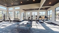 Located at 728 N Church St in Spartanburg, South Carolina, this fitness center is a well-appointed, single-floor facility designed to support a full range of workout routines. The space is defined by its soaring ceilings with a striking dark coffered design accented by recessed lighting, creating a bold yet polished aesthetic. Large floor-to-ceiling windows with black frames line the walls, flooding the interior with natural light and offering open views of the surrounding outdoors. The flooring features durable patterned carpet tiles in shades of blue and gray, providing comfort and style throughout the space. The cardio zone is equipped with treadmills, upright bikes, and a WaterRower rowing machine, all positioned to take advantage of the natural light and views. The strength training area includes a multi-station cable machine with cross-functional attachments, a comprehensive dumbbell rack, weight benches, and additional resistance machines. Full-length mirrors are installed along one wall, enhancing the sense of space and allowing users to monitor their form. A separate glass-paneled room provides an enclosed area for focused training or stretching. The overall ambiance is modern and energizing, combining architectural sophistication with practical fitness functionality.
