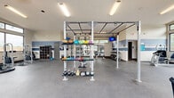 This fitness center occupies a generous single-floor footprint, thoughtfully designed to accommodate a wide range of workout styles and fitness goals. The open-concept layout is organized into distinct functional zones, allowing multiple users to train simultaneously without congestion. Large floor-to-ceiling windows and glass doors line the perimeter walls, flooding the space with natural light and offering views of the surrounding outdoor area, creating an inviting and energizing atmosphere.

The cardio zone features a row of treadmills, elliptical trainers, and a rowing machine, all positioned near the expansive windows. A dedicated strength training area includes a freestanding multi-functional rig equipped with a pull-up bar, along with barbells, benches, and weight plates for compound lifts. A well-stocked dumbbell rack with a flat bench provides options for isolation exercises. An additional section houses cable machines and selectorized strength equipment for targeted training.

Colorful medicine balls and kettlebells in varied weights are neatly organized on dedicated racks, supporting functional fitness and conditioning workouts. Wall-mounted flat-screen televisions are positioned throughout the space for entertainment. Durable rubber flooring covers the entire area, providing safety and comfort. Storage cubbies and wall-mounted accessories round out the practical amenities of this comprehensive fitness facility.