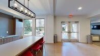 This single-floor amenity space is thoughtfully designed to accommodate a variety of social and recreational activities. The layout is divided into several distinct zones that flow seamlessly into one another. A welcoming kitchenette anchors one end of the space, featuring stainless steel appliances, a full-size refrigerator, tile backsplash, wood cabinetry, and a peninsula counter lined with bold red barstools beneath an industrial-style pendant light fixture. Light wood flooring and large windows overlooking a landscaped outdoor patio with a grill area bring in natural light and a sense of openness.

Adjacent to the kitchenette, a spacious game area with neutral carpet hosts a foosball table and casual lounge seating, with a glass-panel partition wall offering visual separation from a semi-private room beyond. A media lounge at the opposite end of the space features a large wraparound leather sectional, ottomans, a wall-mounted flat-screen television, and textured accent walls, creating an intimate, theater-like atmosphere. Multiple windows with views of greenery add dimension to the darker, moodier palette of this area. Recessed lighting throughout ensures each zone is well-illuminated, while glass double doors provide convenient exterior access.