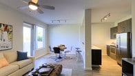 This well-designed single-floor residence offers a clean, contemporary aesthetic throughout its open-concept floor plan. The living area anchors the space with a ceiling fan overhead and flows naturally into a dedicated dining area, where a round table and modern chrome chairs sit beneath track lighting. Large windows along one wall flood the space with abundant natural light, creating a warm and inviting atmosphere that enhances the neutral color palette of white walls and light hardwood-style flooring. A tufted leather ottoman adds a touch of texture and character to the living zone. The dining area transitions smoothly into the kitchen, which is separated by a partial wall and features a sleek dark-cabinetry design with a contrasting light countertop peninsula — ideal for casual seating or meal prep. Additional track lighting in the kitchen ensures the workspace is well-illuminated. The overall ambiance is modern, airy, and functional, with thoughtful architectural details such as clean column accents and an uncluttered open layout that maximizes the sense of space. This residence is well-suited for those who appreciate contemporary design and easy, flowing everyday living.
