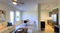 This well-designed single-floor residence offers a clean, contemporary aesthetic throughout its open-concept floor plan. The living area anchors the space with a ceiling fan overhead and flows naturally into a dedicated dining area, where a round table and modern chrome chairs sit beneath track lighting. Large windows along one wall flood the space with abundant natural light, creating a warm and inviting atmosphere that enhances the neutral color palette of white walls and light hardwood-style flooring. A tufted leather ottoman adds a touch of texture and character to the living zone. The dining area transitions smoothly into the kitchen, which is separated by a partial wall and features a sleek dark-cabinetry design with a contrasting light countertop peninsula — ideal for casual seating or meal prep. Additional track lighting in the kitchen ensures the workspace is well-illuminated. The overall ambiance is modern, airy, and functional, with thoughtful architectural details such as clean column accents and an uncluttered open layout that maximizes the sense of space. This residence is well-suited for those who appreciate contemporary design and easy, flowing everyday living.