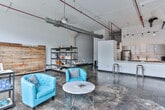This single-floor industrial loft space masterfully blends raw, historic character with modern amenities. The main open area commands attention with its soaring vaulted ceilings, exposed HVAC ductwork, and red fire suppression piping that trace the roofline overhead. Polished concrete floors with natural patina run throughout, reinforcing the building's authentic industrial heritage. Original masonry walls, partially exposed, add texture and depth alongside freshly painted white surfaces that brighten the interior.

Large, steel-framed factory-style windows line the perimeter, drawing in generous amounts of natural light and offering views of the surrounding environment. The open main room is versatile and expansive, currently appointed with a casual lounge area featuring vibrant teal accent chairs, a round marble-top coffee table, freestanding metal shelving units, and a reclaimed wood accent wall.

A compact but well-equipped kitchenette features sleek gray cabinetry, white quartz countertops, subway tile backsplash, and stainless steel appliances. A private restroom includes a modern vanity with a dark stone countertop, undermount sink, large mirror, and contemporary lighting. A corridor with a wood-paneled door connects the service areas to the open main floor, completing a functional and thoughtfully designed layout.