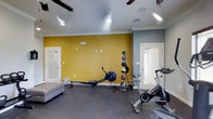This fitness center offers a dedicated space for a full range of workout activities, thoughtfully designed to support both cardio and strength training. The room immediately draws attention with its bold yellow accent wall, which adds an energizing and motivating ambiance to the space. The remaining walls are finished in a cool gray tone, creating a balanced and modern aesthetic throughout.

The flooring is covered in durable rubberized material, providing both safety and comfort during exercise. Equipment on hand includes a rowing machine for full-body cardio conditioning, a spinning bike ideal for low-impact cycling workouts, and a vertical medicine ball rack stocked with multiple weighted balls for functional strength and conditioning exercises. A padded bench is also available, offering additional versatility for stretching or workout routines.

Overhead, ceiling fans help maintain airflow and comfort during intense sessions, complemented by recessed lighting that ensures the space is well-lit. Multiple doors provide convenient access points, enhancing the overall flow of the facility. Crown molding adds a subtle architectural polish to the room. This fitness center is a practical and well-appointed facility designed to support an active and healthy lifestyle.