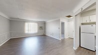 This well-appointed single-floor unit presents a bright, neutral palette and thoughtfully updated finishes throughout. The spacious main living area is a standout, showcasing smooth hardwood-style flooring that reflects natural light filtering in through large vertical-blind-draped windows. Decorative wainscoting panels line the lower walls, adding a refined architectural detail that elevates the overall design. The open layout connects seamlessly to additional rooms, creating a comfortable flow throughout the home.

The kitchen is outfitted with crisp white cabinetry, brushed nickel hardware, and generous marble-look countertops that wrap around the corner, providing ample workspace. A stainless steel sink with a modern pull-down faucet completes the functional yet stylish cooking space.

The bedrooms offer plush carpeting in soft neutral tones, with natural light entering through double-hung windows fitted with blinds. These rooms provide a comfortable and quiet retreat. A secondary living or flex room mirrors the wainscoting detail seen in the main area, offering additional versatile space with the same warm hardwood-style flooring.

The bathroom is clean and efficiently designed, featuring a full soaking tub and shower combination, a toilet, vanity, and wood-look flooring, all finished in bright white tones that enhance the sense of cleanliness and space.