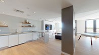 This single-floor space showcases a cohesive modern design marked by clean lines, a neutral white-and-gray palette, and light hardwood flooring that flows throughout. The open-plan kitchen is outfitted with flat-front white cabinetry, stainless steel countertops, a stainless side-by-side refrigerator, a built-in microwave, and a stainless sink. Warm-toned floating wood shelves add contrast against the white walls, while a peninsula with bar seating connects the kitchen to the main living area. The living space centers on a sleek built-in wall unit housing a large flat-screen TV, flanked by white gloss cabinetry. A sectional sofa, mid-century coffee table, and patterned area rug create a comfortable seating arrangement, while floor-to-ceiling sliding glass doors open to an outdoor terrace with pool and city views, flooding the room with natural light. A dedicated game room features a contemporary ping pong table set against a bold geometric blue accent wall mural, with direct access to the outdoor terrace. Architectural ceiling details, recessed lighting, and a gray structural column add visual interest throughout the space, reinforcing its modern, design-forward character.