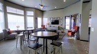 This single-floor communal space is thoughtfully designed to accommodate both casual gatherings and quiet relaxation. The room features notably high ceilings accentuated by crown molding and recessed lighting, contributing to an open, airy atmosphere. A ceiling fan with a light fixture anchors the center of the room, providing both functionality and a subtle design element.

The layout is divided into distinct seating zones. A high-top bistro table with distressed metal stools occupies the foreground, offering a casual dining or social area. Further into the room, a lounge zone is arranged around a flat-screen television mounted above a white media console, flanked by a dark leather sectional sofa and vibrant red accent chairs that add a pop of color. A patterned area rug grounds this seating arrangement.

The walls are adorned with an eclectic gallery of framed artwork, adding personality to the space. Large windows fitted with blinds allow generous natural light to filter in, while a glass door on one side provides views to an outdoor area. The tile flooring throughout is both durable and visually cohesive, tying the entire space together.
