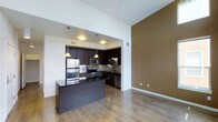 Located at 10400 East 29th Drive in Denver's vibrant Central Park neighborhood, this single-floor residence offers a thoughtfully designed open layout that blends modern style with functional living. The kitchen is a standout feature, showcasing rich dark cabinetry, granite countertops, and a full suite of stainless steel appliances including a refrigerator, range, and built-in microwave. A mosaic tile backsplash adds texture and visual interest, while pendant lighting suspended from the ceiling creates a warm, inviting atmosphere over the breakfast bar peninsula — ideal for casual dining or entertaining. The kitchen flows seamlessly into the main living area, which is highlighted by warm hardwood-style flooring that extends throughout the space, contributing to a cohesive and contemporary aesthetic. An accent wall in a deep earth tone grounds the living area and adds a layer of design sophistication. The generous ceiling height enhances the overall sense of openness and volume. Clean white walls contrast beautifully with the darker design elements, creating a balanced and modern interior that is both welcoming and stylish.