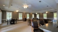 This single-floor space presents a well-appointed interior designed for comfortable gathering and socializing. The room is finished with warm, taupe-toned walls accented by crisp white crown molding and window casings, creating a refined yet welcoming atmosphere. Multiple ceiling fans with light fixtures are evenly distributed throughout, ensuring both comfort and consistent illumination across the generous open layout.

Natural light flows in through numerous double-hung windows dressed with white plantation shutters, offering views of the surrounding greenery while maintaining privacy. The neutral carpeted flooring complements the earthy wall tones and ties the space together cohesively.

The furnishings are arranged to accommodate various uses, with leather and ornate wood-framed seating positioned around granite-topped tables in the foreground, and additional dining-height table-and-chair sets visible toward the rear. Decorative accents, including table lamps and vases, add warmth and character throughout. Recessed lighting supplements the ceiling fans, providing layered illumination. Overall, the space combines classic architectural details with functional, multi-purpose furnishings to create an inviting communal environment.