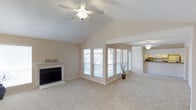 This single-floor home in Arlington offers a welcoming and open interior layout designed for comfortable everyday living. The spacious living room immediately draws the eye upward with its dramatic vaulted ceiling, which adds a sense of grandeur and airiness to the space. A ceiling fan is centrally mounted overhead, contributing both style and practicality. The room is anchored by a classic fireplace with a clean white surround, providing a warm and inviting focal point. Large windows along the wall flood the living area with generous natural light, offering views of the surrounding landscape while keeping the interior bright and airy. Soft, neutral-toned carpeting extends throughout, complementing the light beige walls and creating a calm, cohesive aesthetic. The open floor plan flows naturally from the living room into the kitchen, which is visible in the background and features warm cabinetry and overhead lighting. This seamless connection between living and kitchen spaces makes the layout ideal for entertaining and day-to-day functionality. The overall ambiance is clean, bright, and inviting, with thoughtful architectural details that elevate the character of the home.