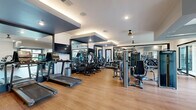 Located at 2880 Avila Cross Cir in Oviedo, Florida, this fitness center offers a thoughtfully designed exercise space that balances functionality with modern aesthetics. The single-floor layout provides a generously open workout area that feels spacious and well-organized.

The exercise room features warm wood-look flooring throughout, creating an inviting atmosphere that complements the contemporary design. Strategically placed large mirrors along the walls visually expand the space and allow users to monitor their form during workouts. Recessed lighting and ceiling fans maintain a bright, comfortable environment, while bold dark-framed architectural ceiling details add a modern, polished touch.

The space is comprehensively equipped with a full lineup of Matrix-brand cardio machines, including treadmills, ellipticals, and stair climbers, arranged in a dedicated row. The strength training area features multiple cable and selectorized resistance machines, a functional training station, adjustable benches, and a dumbbell rack. Large windows and glass doors along the perimeter flood the interior with natural light and offer views of the surrounding outdoor greenery.

A built-in shelving unit and smart control panel are also present, enhancing the room's usability. The overall ambiance is clean, modern, and energizing, making this fitness center a well-appointed facility for a comprehensive workout experience.