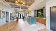 Located at 7800 District Crossing Dr in Sunbury, Ohio, this thoughtfully designed leasing office offers a polished and welcoming first impression. The single-floor space is organized into distinct functional zones that flow seamlessly into one another.

Upon entry, visitors are greeted by a striking reception area anchored by a teal geometric-tiled front desk, illuminated by elegant gold pendant lights and a glamorous chandelier overhead. The coffered ceiling features a dramatic marbled mural, adding a sophisticated artistic touch. A living wall bearing a glowing "Welcome Home" sign is set against navy cabinetry, reinforcing the warm, community-focused atmosphere.

Adjacent to the reception, a bright dining area with wood-toned chairs and large glass doors offers views of the outdoor pool area, allowing ample natural light to fill the space. Light wood-look flooring and large-format tile run throughout, unifying the open layout.

A separate lounge area features soaring vaulted ceilings with exposed wood beam trusses, a cozy fireplace with a wall-mounted television above a natural wood mantel, and comfortable seating. A private nook with classic wainscoting, a leather armchair, and natural light provides a quiet space for one-on-one conversations. Curated artwork and textured wall treatments complete the refined, community-centered design.