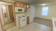 This two-floor space offers a practical and comfortable layout designed for everyday living. The kitchen is equipped with warm-toned oak cabinetry, providing ample storage overhead and at counter level. White appliances, including a coil-burner electric range, an over-the-range microwave, and a full-size refrigerator, are all included, making the kitchen move-in ready. The flooring in the kitchen features a neutral tile pattern that is easy to maintain. The kitchen opens directly into an adjacent living area, which is finished with wall-to-wall carpet and benefits from a window that allows natural light to filter in, creating a bright and welcoming atmosphere. The textured ceiling is consistent throughout the main level, giving the space a classic, traditional feel. The second floor expands the overall footprint of the home, offering additional space for sleeping quarters or flexible use. The overall layout is straightforward and efficient, with a neutral color palette that provides a versatile backdrop for a variety of interior styles. The connection between the kitchen and living areas supports an open, easy flow for daily activities.