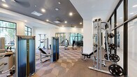 This modern fitness center offers a thoughtfully designed, single-floor workout facility that provides ample space for a comprehensive exercise experience. The interior is defined by crisp white walls accented by a bold dark-painted ceiling in the central zone, creating a dynamic visual contrast. Recessed lighting and ceiling fans throughout ensure a bright and comfortable environment during workouts.

The facility is outfitted with a full suite of Matrix-brand strength training machines, adjustable benches, a cable crossover system, a dumbbell rack, and a variety of free weights. Stability balls and kettlebells are also available, supporting functional and accessory training. The floor features a distinctive geometric, diamond-patterned carpet that adds texture and visual interest to the space.

Large floor-to-ceiling windows and glass-panel framed doors line one side of the facility, flooding the space with natural light and offering views of an outdoor courtyard and pool area. Mirrored walls further amplify the sense of openness and allow users to monitor their form during exercise. The entry is marked by sleek black-framed glass partition doors, adding a refined, contemporary aesthetic. Overall, the fitness center balances functionality with modern design, creating an inviting and well-appointed environment for a full range of strength and fitness training activities.
