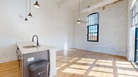 Located at 612 W Main Ave in downtown Gastonia, NC, this single-floor loft-style unit seamlessly blends industrial heritage with modern renovations. The open living area is defined by whitewashed exposed brick walls, soaring ceilings with a tongue-and-groove wood ceiling detail, and oversized arched and grid-pane windows that flood the space with abundant natural light and offer street-level views of the surrounding cityscape. Ceiling fans and stylish pendant lighting add both function and character throughout.

Warm-toned hardwood floors run continuously throughout the unit, unifying every room with a clean, cohesive aesthetic. The kitchen features a central island with a light countertop, an undermount sink with a matte black faucet, pendant lighting above, and stainless steel appliances, all positioned to overlook the expansive open living and dining area.

The bedrooms are bright and well-proportioned, with clean white walls, hardwood flooring, and closets outfitted with built-in shelving and hanging rods. The bathroom is finished with hardwood flooring, a full tub-shower combination, matte black fixtures, and clean white surfaces throughout. Industrial ductwork and exposed structural elements contribute to the authentic loft character of this thoughtfully updated space.