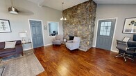 This single-story leasing center in Homewood, Alabama, offers a welcoming and well-appointed environment designed to make visitors feel at ease. The main reception area is expansive and open, characterized by soaring vaulted ceilings with exposed beams and rich, warm hardwood flooring that extends throughout the entire space. Pendant lighting fixtures hang from the high ceilings, complementing table lamps to create a balanced, inviting atmosphere.

A dramatic floor-to-ceiling stone fireplace serves as the architectural centerpiece of the space, flanked by a pair of upholstered accent chairs that provide a comfortable seating vignette. Opposite this area, a lounge seating arrangement features a dark upholstered sofa, a glass-top coffee table, and a patterned area rug, offering a relaxed setting for consultations.

The entry area is accessed through glass-paneled double doors, framed by striped sheer curtains that allow natural light to filter into the space. A branded welcome mat greets visitors upon arrival. Additional functional areas include a workstation with a desk and task seating, as well as a service counter. A private office is accessible through interior doorways, providing space for one-on-one leasing conversations.