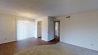 This single-floor residence offers a straightforward and functional layout with clearly defined living spaces. The main living area features a combination of plush carpeting and dark hardwood-style flooring, creating a natural visual transition between zones. A classic chandelier hangs overhead, providing a traditional decorative touch, while a textured ceiling adds subtle dimension to the space.

Vertical blinds cover a sliding glass door along one wall, allowing for adjustable natural light and access to what appears to be an outdoor area. The neutral white walls throughout create a clean, bright atmosphere that lends itself well to a variety of interior styles.

From the living area, an open doorway leads directly into the kitchen, which is equipped with a white refrigerator and appears to include cabinetry with ample storage. A second doorway reveals an adjacent room, likely a bedroom or bathroom, featuring white cabinetry or a vanity, suggesting practical utility. The HVAC vent on the upper wall indicates a central air system serving the space. Overall, the residence presents a simple, well-connected layout suited for comfortable everyday living.
