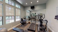 Located at 973 Westcreek Ln in Thousand Oaks, CA, this single-floor fitness center is thoughtfully designed to support a well-rounded workout experience. The space is finished with neutral sage-green walls, recessed lighting, and durable carpet-style flooring, creating a clean and functional atmosphere throughout.

The cardio area is well-equipped, featuring a treadmill, an upright elliptical trainer, and a recumbent stationary bike, all positioned along a wall of large multi-pane windows that frame views of surrounding mature trees. A wall-mounted television is positioned above a full-length mirror, adding both entertainment and a sense of expanded space.

The strength training section includes a multi-function cable machine suitable for a wide range of resistance exercises, a complete rack of hex dumbbells, a stability ball, and a flat utility bench. Large mirrors line the walls, enhancing the openness of the room and allowing users to monitor their form during workouts.

Entry to the fitness center is through elegant white French doors with glass panes, which connect to an adjacent community space visible beyond. Sanitation stations and posted facility guidelines are conveniently mounted near the entrance, underscoring the center's commitment to cleanliness and user comfort. Natural light pours in from multiple window elevations, giving the space a bright and welcoming ambiance.