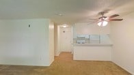 This single-floor residence presents a straightforward and functional layout designed for comfortable everyday living. The main living area is open and bright, finished with wall-to-wall carpeting that adds warmth underfoot. A ceiling fan with an integrated light fixture is centrally positioned, providing both air circulation and ambient lighting throughout the space.

The kitchen is situated in an open-plan arrangement adjacent to the living area, separated by a raised breakfast bar or pass-through counter. White cabinetry lines the kitchen walls, complemented by white appliances including a range and a full-sized refrigerator. The kitchen flooring transitions to a hard surface, offering a practical contrast to the carpeted living space.

A hallway leads from the main living area toward the entry door, which is visible at the far end and features overhead lighting. The textured popcorn ceiling is consistent throughout, and the clean white walls contribute to a bright, neutral interior palette. The overall ambiance is understated and move-in ready, with a practical design that makes efficient use of the available space across the single level.