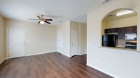 This single-floor space showcases a well-designed open layout that connects the main living area to the kitchen through a distinctive arched pass-through opening. The living area is finished with rich, dark hardwood flooring that runs throughout, providing a warm and grounding contrast to the neutral beige walls. A ceiling fan with a built-in light fixture is centrally mounted, contributing both to air circulation and ambient lighting. The walls are painted in a soft, neutral tone that enhances the brightness of the space and complements the clean white trim and baseboards. Multiple doors are visible leading to additional rooms or storage areas, suggesting functional versatility. The arched pass-through connects to the kitchen, where a black refrigerator is partially visible, hinting at modern appliances within. The layout is open and unobstructed, allowing for flexible furniture arrangements. HVAC vents are positioned in the ceiling, indicating a centralized climate control system throughout the unit. Overall, the space presents a clean, move-in ready aesthetic with a balance of practical design and inviting warmth.