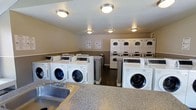 This shared laundry facility is thoughtfully designed to accommodate a high volume of laundry needs in a clean, organized environment. The room is outfitted with a row of front-loading washing machines positioned along the floor, complemented by multiple stacked dryer units lining the back wall, providing ample capacity for simultaneous use. The machines are modern, commercial-grade appliances well-suited for frequent, heavy-duty operation. A granite-style folding counter runs along one side of the room, offering a convenient surface for sorting and folding laundry, with a utility sink integrated into the countertop for added functionality. Instructional signage is posted on the walls, ensuring users can operate the equipment with ease. The ceiling features evenly distributed flush-mount light fixtures that illuminate the space with bright, consistent lighting. The room is finished with neutral-toned walls and durable flooring, creating a tidy and practical atmosphere. Overall, this laundry room is a functional, well-maintained communal space designed to serve residents efficiently.
