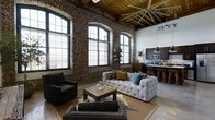 This single-floor space makes an immediate impression with its rich industrial character and thoughtful design. Exposed brick walls stretch toward soaring ceilings supported by warm wooden beams, creating a dramatic backdrop throughout the open-concept interior. A large ceiling fan anchors the living area, which is bathed in abundant natural light pouring through a series of tall, arched windows fitted with dark frames — a defining architectural feature that adds both grandeur and historic charm.

The living area is generously proportioned, accommodating a tufted Chesterfield-style sofa, a rustic wooden coffee table, and additional seating arranged around a cowhide-accented area rug. The eclectic furnishings complement the raw materiality of the space, balancing industrial elements with comfort and warmth.

Seamlessly connected to the living area, the kitchen features dark cabinetry, stainless steel appliances, and a raised bar-height counter with seating — ideal for casual dining or entertaining. Pendant lighting over the kitchen adds a refined touch against the otherwise rugged aesthetic.

The overall ambiance is bold yet inviting, merging the character of a historic industrial building with modern livability. Exposed brick, natural wood, soaring ceilings, and ample light combine to create a space that is both visually compelling and highly functional.