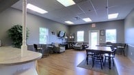 This single-floor space is thoughtfully arranged across two connected zones, each serving distinct functional purposes while maintaining a cohesive aesthetic throughout. Warm hardwood flooring runs continuously across both areas, tying the layout together and contributing to an inviting, comfortable atmosphere.

The first zone presents an open, lounge-style layout with gray-toned walls and a drop ceiling fitted with recessed panel lighting that provides consistent, even illumination. Multiple windows along the walls allow natural light to filter in, offering views of surrounding greenery. The furnishings include a mix of armchairs, a bar-height table with stools, a dining-style table with chairs, and a small media setup along one wall, making the space adaptable for social gatherings, informal meetings, or relaxed seating arrangements.

The second connected zone continues the hardwood flooring and features a large, wraparound sectional sofa as its centerpiece, complemented by additional seating and potted plants that introduce a touch of natural warmth. This area feels more lounge-oriented and provides a comfortable, casual setting. Together, both zones form a well-balanced, multi-purpose space designed to accommodate a variety of activities in a welcoming environment.
