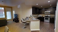 This single-floor space showcases an open-concept layout that integrates the kitchen and living areas into one cohesive environment. The kitchen is a standout feature, outfitted with rich, dark-toned cabinetry that extends to the upper walls, providing ample storage. Granite-style countertops add a touch of elegance, while stainless steel appliances, including a range and microwave, offer modern functionality. A double-basin sink is positioned along the peninsula, which also serves as a breakfast bar with stylish swivel bar stools — ideal for casual dining or conversation.

The flooring transitions from warm hardwood-style planks in the kitchen to plush carpet in the adjoining living area, subtly defining each zone within the open floor plan. Neutral, warm-toned walls create a welcoming ambiance throughout, complemented by recessed lighting that ensures the space is well-lit. The living area accommodates a flat-screen television setup, making it a comfortable space for relaxation. Natural light filters in through a window near the living area, enhancing the bright and airy feel of the overall space.