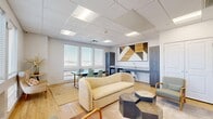 Located at 1977 S 300 W in Salt Lake City, Utah, this single-floor living space offers a thoughtfully designed and multifunctional layout. The room is lined with a generous bank of large windows fitted with white horizontal blinds, allowing ample natural light to pour in while offering views of the surrounding cityscape. Light hardwood flooring runs throughout, complementing the bright white walls and drop-panel ceiling with recessed fluorescent lighting.

The living area is furnished with a plush cream sofa, a wood-framed green accent chair, and a set of geometric hexagonal coffee tables arranged on a neutral area rug, creating a comfortable and stylish seating zone. A wall-mounted flat-screen television anchors one side of the room, while a striking geometric wood-panel accent wall serves as a bold focal point above a built-in kitchenette. The kitchenette includes a countertop with a sink, microwave, and refrigerator, adding practical utility to the space.

A dining area with a dark rectangular table and teal chairs sits adjacent to the windows, ideal for meetings or shared meals. Additional seating is provided by a patterned armchair and side table tucked into a corner. A wooden credenza with a wall-mounted sink rounds out the room's functional amenities, making this a versatile and well-equipped space.