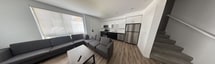 This contemporary 840 square foot apartment offers a thoughtful layout with 3 bedrooms and 2 bathrooms. The heart of the home is an open-concept living area that seamlessly integrates the kitchen and living room. The kitchen boasts a striking contrast of white upper cabinets and dark lower cabinets, complemented by stainless steel appliances and a subway tile backsplash. Wood-look flooring runs throughout the main living areas, adding warmth to the modern aesthetic.

The primary bedroom, measuring 140 square feet, features carpeting and a large window that allows ample natural light. All bedrooms are equipped with closet space and offer versatility for various uses. The bathrooms showcase a clean, white design with tiled surrounds in the shower/tub combinations.

The living room is furnished with comfortable grey seating and a TV area, perfect for relaxation or entertainment. Recessed lighting throughout the apartment provides a sleek, unobtrusive illumination solution. Large windows in the living area and bedrooms not only brighten the space but also offer views of the surrounding area.

This single-story dwelling combines functionality with style, making efficient use of its compact footprint while maintaining a sense of spaciousness through its thoughtful design and light color palette.