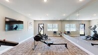 Located at 801 Booth Rd in Warner Robins, Georgia, this fitness center offers a clean, functional workout space designed to support a variety of exercise routines. The single-floor layout is finished with neutral gray walls, white baseboards, and patterned carpet flooring that provides comfort and durability underfoot. Recessed lighting throughout the ceiling ensures consistent, even illumination across the entire space.

A standout feature is the large floor-to-ceiling mirror spanning one wall, which amplifies the sense of openness and allows users to monitor their form during workouts. The room is equipped with a range of cardio machines, including a treadmill, an elliptical trainer, and a rowing machine, catering to diverse fitness preferences. A dumbbell rack with a tiered set of free weights adds strength-training capability, while medicine balls along the mirrored wall provide additional options for functional fitness.

Multiple windows fitted with horizontal blinds allow natural light to filter in while maintaining privacy, and views of the surrounding community grounds are visible through the glass-paneled exterior door. Decorative framed artwork and a large wall clock add an inviting, polished aesthetic to the space. Seating chairs positioned near the windows offer a convenient rest area. The overall ambiance is bright, tidy, and purposefully designed for an effective and comfortable fitness experience.