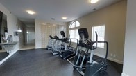 This fitness center is a dedicated, single-floor space designed to support an active lifestyle. The room is outfitted with a lineup of modern cardio equipment, including treadmills and elliptical trainers from reputable brands, all arranged to maximize usability and floor space. The flooring consists of durable rubber matting, providing both safety and comfort during workouts.

Natural light fills the space through multiple windows fitted with blinds, including a distinctive arched window that adds an architectural touch to the room. A decorative glass block wall panel serves as a standout design feature, diffusing soft light while adding visual interest and texture to the interior. Recessed and flush-mount ceiling lights supplement the natural light, ensuring the space is well-illuminated at all times.

The neutral-toned walls and clean layout create an open, focused atmosphere that is both functional and inviting. Ventilation is provided through ceiling vents, contributing to a comfortable environment during exercise. The overall design balances practical fitness facility requirements with thoughtful architectural details, making this space a well-rounded and pleasant environment for physical activity.