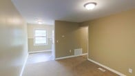 This single-floor space presents a straightforward, functional layout with a neutral color palette throughout. The main living area features wall-to-wall carpeting in a warm beige tone, complemented by crisp white baseboards that frame the room cleanly. Walls are painted in a soft tan hue, creating a cohesive and understated aesthetic.

Flush-mount ceiling light fixtures provide consistent overhead lighting across the space. A doorway opening connects the carpeted main area to an adjoining room finished with light-toned tile flooring, which appears to serve as a utility or transitional space. That secondary area benefits from natural light courtesy of a double-hung window, brightening the interior noticeably.

Floor-level vents are present in both areas, indicating a forced-air HVAC system throughout. A wall-mounted thermostat is visible, offering climate control convenience. The clean, uncluttered condition of the space allows for easy assessment of its layout and dimensions. White trim detailing on doors and baseboards ties the rooms together visually, while the open doorway between areas promotes a connected flow. Overall, the space presents a practical, well-maintained interior ready for occupancy.