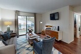 This single-floor residence offers a thoughtfully designed layout encompassing a living area, dining space, galley-style kitchen, two bedrooms, and at least one full bathroom. The living room serves as the central gathering space, featuring rich hardwood-style flooring, a bold geometric-patterned area rug, and a large sliding glass door that floods the room with natural light and opens onto a private balcony with outdoor views. A wall-mounted television and a mid-century modern-style credenza anchor the media wall.

Adjacent to the living area, the dining space accommodates a round table and seating for several, with a pass-through counter connecting directly to the kitchen. The galley kitchen is efficiently organized with gray cabinetry, marble-style countertops, a stainless steel sink, and black appliances including a refrigerator and electric range.

Both bedrooms are carpeted and well-sized, each offering natural light through blinds-fitted windows. One bedroom includes a ceiling fan with integrated lighting. The bathroom features wood-style flooring, a marble-style vanity countertop with an undermount sink, and a tiled shower-tub combination. The overall aesthetic blends contemporary and mid-century modern influences throughout.