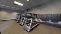 This single-level fitness center is thoughtfully designed to accommodate a wide range of workout needs within a well-organized layout. The space is divided into distinct functional zones, each tailored to a specific type of exercise. The cardio area is equipped with multiple treadmills and elliptical machines, all positioned in front of a dramatic, large-scale mural depicting a Burlington steam locomotive — a bold artistic feature that adds character and visual energy to the room.

Adjacent to the cardio zone, the strength training area includes a multi-station cable machine, a dumbbell rack, adjustable benches, and a recumbent bike, providing ample options for resistance training. A dedicated stretching and floor exercise zone features rubber matting, a full-length mirror wall, and a wall-mounted ballet barre, making it suitable for flexibility work, yoga, or dance-inspired fitness routines.

The interior finishes are clean and functional, with neutral-toned carpeting, drop tile ceilings with recessed and fluorescent lighting, and gray-painted walls. Windows along one side allow natural light to filter into the space, contributing to an open, energizing atmosphere well-suited for daily fitness activity.