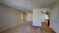 This single-floor unit is organized around an open-concept layout that connects the main living area to the kitchen via a central partial-wall divider. The living area is finished with wall-to-wall beige carpet, creating a warm and comfortable feel underfoot. Neutral beige walls and white trim run throughout the space, contributing to a clean, understated aesthetic. A textured popcorn ceiling spans the entirety of the unit, and track lighting is mounted overhead in the living area, providing adjustable illumination.

The kitchen is distinctly separated by the transition from carpet to hardwood-style flooring, offering a practical and visually defined cooking zone. Dark wood cabinetry lines the kitchen walls, providing ample storage, while white appliances — including a refrigerator, range, and overhead range hood — offer a classic contrast. Fluorescent overhead lighting illuminates the kitchen efficiently.

A doorway near the entry leads to additional rooms, and a secondary doorway visible from the living area suggests access to a bathroom or additional space. The overall ambiance of the unit is functional and straightforward, with a neutral palette that offers flexibility for personalization.