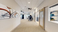 Located at 3483 SW 45th St in Gainesville, FL, this single-story fitness facility offers a thoughtfully designed layout across a generous floor area, accommodating a comprehensive range of workout needs. The space is divided into three distinct zones, each tailored to a specific training focus.

The main strength training area features plush carpet flooring and houses an extensive collection of selectorized resistance machines, cable systems, and free weights, including a full dumbbell rack. Ceiling fans and recessed lighting keep the environment comfortable and well-lit, while large windows with outdoor views bring in natural light. Vibrant athletic-themed artwork adorns the walls, adding an energetic ambiance. A wall-mounted TV, water fountain station, and dedicated entry doors provide practical amenities.

The cardio zone is lined with windows that flood the space with daylight, and is equipped with treadmills, an elliptical, and a stationary bike, all positioned to take advantage of the natural light and outdoor scenery.

The group exercise studio stands out with its light hardwood flooring, full-length wall mirrors, and a unique glass-panel garage door that opens directly to an outdoor green space. The studio is stocked with yoga mats, stability balls, exercise bands, and dumbbells, making it ideal for group classes or individual training sessions.