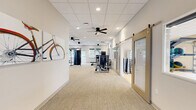 Located at 3483 SW 45th St in Gainesville, FL, this single-story fitness facility offers a thoughtfully designed layout across a generous floor area, accommodating a comprehensive range of workout needs. The space is divided into three distinct zones, each tailored to a specific training focus.

The main strength training area features plush carpet flooring and houses an extensive collection of selectorized resistance machines, cable systems, and free weights, including a full dumbbell rack. Ceiling fans and recessed lighting keep the environment comfortable and well-lit, while large windows with outdoor views bring in natural light. Vibrant athletic-themed artwork adorns the walls, adding an energetic ambiance. A wall-mounted TV, water fountain station, and dedicated entry doors provide practical amenities.

The cardio zone is lined with windows that flood the space with daylight, and is equipped with treadmills, an elliptical, and a stationary bike, all positioned to take advantage of the natural light and outdoor scenery.

The group exercise studio stands out with its light hardwood flooring, full-length wall mirrors, and a unique glass-panel garage door that opens directly to an outdoor green space. The studio is stocked with yoga mats, stability balls, exercise bands, and dumbbells, making it ideal for group classes or individual training sessions.