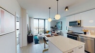 This single-floor residence is designed with a clean, contemporary aesthetic throughout. The open-concept layout seamlessly connects the kitchen, dining, and living areas, creating a cohesive and functional living space. Light wood-tone flooring runs throughout, complementing the bright white walls and ceilings.

The kitchen is outfitted with flat-front white cabinetry extending to the ceiling, a textured tile backsplash, and a full suite of stainless steel appliances including a range, dishwasher, microwave, and refrigerator. A central island with a quartz countertop provides additional prep space and casual seating for bar-height chairs. Globe pendant lights hang above the island, adding a modern, warm accent.

The living area opens to floor-to-ceiling windows with sheer draping curtains, offering expansive urban views and filling the space with natural light. The bedroom continues the city view theme, featuring large windows with access to a private balcony, soft blue accent walls, and ample room for a sitting area.

The bathroom features dark textured tile surrounding a full soaking tub and shower combination, contrasted by a white floating vanity, large mirror, and warm globe lighting. The overall ambiance is bright, modern, and urban.