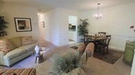 This single-floor residence offers a welcoming and cohesive layout that blends comfort with functionality. The living area is generously proportioned, featuring soft wall-to-wall carpeting that adds warmth underfoot and neutral-toned walls that create a bright, airy atmosphere. A stylish glass-top coffee table and striped upholstered seating anchor the living space, complemented by decorative greenery that brings a touch of nature indoors. Adjacent to the living area, the dining space is defined by an elegant chandelier with candelabra-style lighting, casting a warm glow over a round wooden dining table surrounded by classic cross-back chairs. A patterned area rug beneath the dining set adds texture and visual interest. The kitchen is visible through an open passageway, featuring white cabinetry, light countertops, and tile flooring that contrasts with the carpeted living areas. A long hallway extends toward additional rooms, suggesting a thoughtful floor plan that provides both open communal spaces and private retreats. The overall ambiance is clean, inviting, and well-appointed, making it a comfortable and practical living environment.