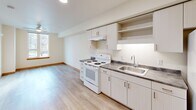 Located at 12370 SE Ash St in Portland, Oregon, this efficiently designed one-bedroom, one-bathroom unit offers a thoughtful layout across a single floor. The open-plan living, dining, and kitchen area forms the heart of the home, featuring light wood laminate flooring, crisp white walls, and recessed lighting that create a bright, clean aesthetic throughout. A ceiling fan adds comfort to the living area, which receives natural light through a street-facing window.

The kitchen is well-appointed with light gray cabinetry offering ample upper and lower storage, open display shelving, a dark laminate countertop, a stainless steel sink, an electric range, and a full-size refrigerator — all arranged along a single efficient wall.

The bedroom is a cozy, sun-lit room with a window overlooking greenery, light wood floors, and warm wood baseboards. It connects directly to a generously sized walk-in closet outfitted with wire shelving and hanging rods, providing excellent storage capacity relative to the unit's overall footprint.

The bathroom features a single-sink vanity with dark countertop, white cabinetry, a toilet, and a full shower/tub combination, all finished in a clean, neutral palette. Natural wood-trimmed doors throughout the unit add a warm, distinctive touch to the overall design.