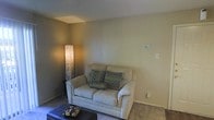 This single-floor space features a modestly sized living area characterized by neutral, earthy tones throughout. The room is finished with wall-to-wall carpeting in a warm beige shade, contributing to a comfortable and casual atmosphere. The walls are painted in a soft, muted green-gray tone, complemented by a textured ceiling that adds subtle dimension to the space.

The living area is anchored by a cream-colored loveseat adorned with decorative patterned throw pillows in muted blue and gray tones. A sleek, dark ottoman serves as a coffee table, adding a functional and contemporary contrast to the lighter furnishings. Ambient lighting is provided by a tall, latticed floor lamp positioned in the corner, casting a warm, soft glow across the room.

The space features a wall-mounted thermostat or control panel, and standard electrical outlets are conveniently placed along the walls. The overall design is straightforward and functional, with a clean, uncluttered layout that makes efficient use of the available space. The neutral color palette and soft lighting create a relaxed, inviting ambiance suitable for everyday living.